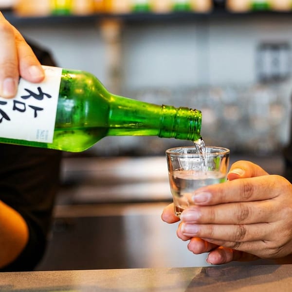 Two people's hands pouring Soju from a green bottle into a shot glass.