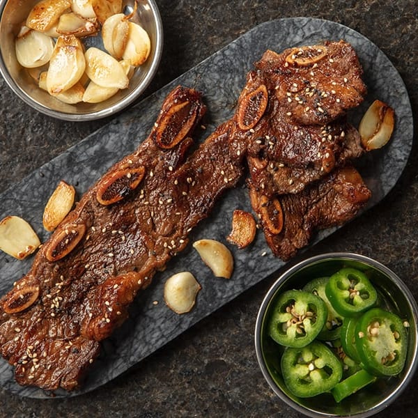 Korean BBQ short ribs with roasted garlic and sliced jalapeños on a stone platter.